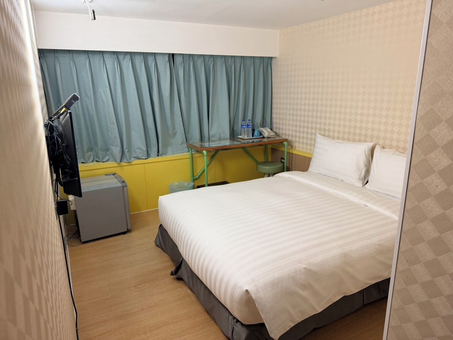 A hotel room with a bed, a desk, a chair, a small table, a telephone, and a window with curtains.