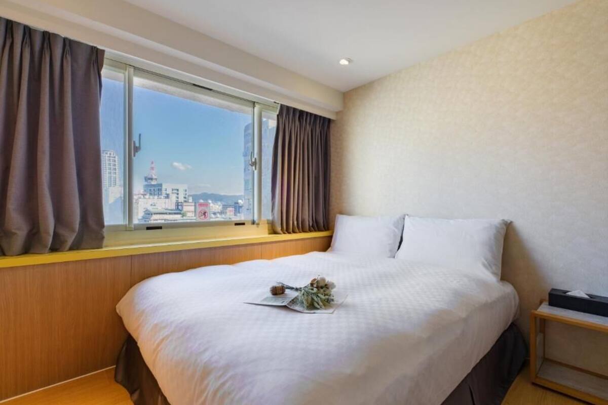 A hotel room with a large bed, a window with curtains, a wooden headboard, and a bedside table with a lamp.