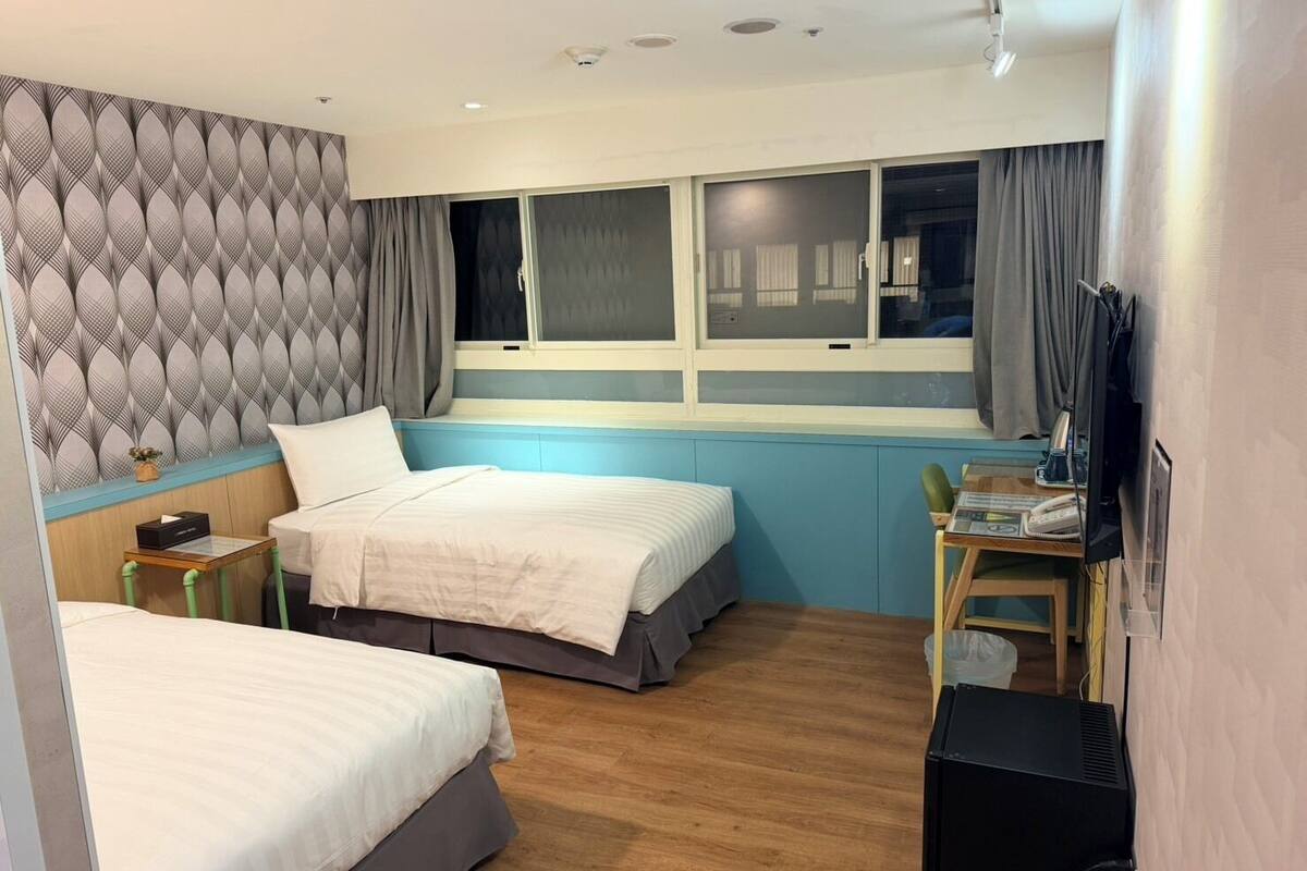 A hotel room with two beds, a desk with a chair, a TV, and a window with curtains.
