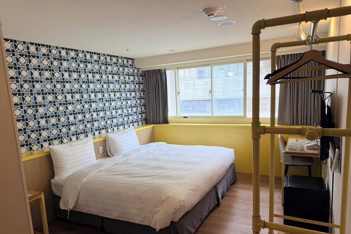 A hotel room with a bed, a window with curtains, a wall with a geometric pattern, and a wooden floor.