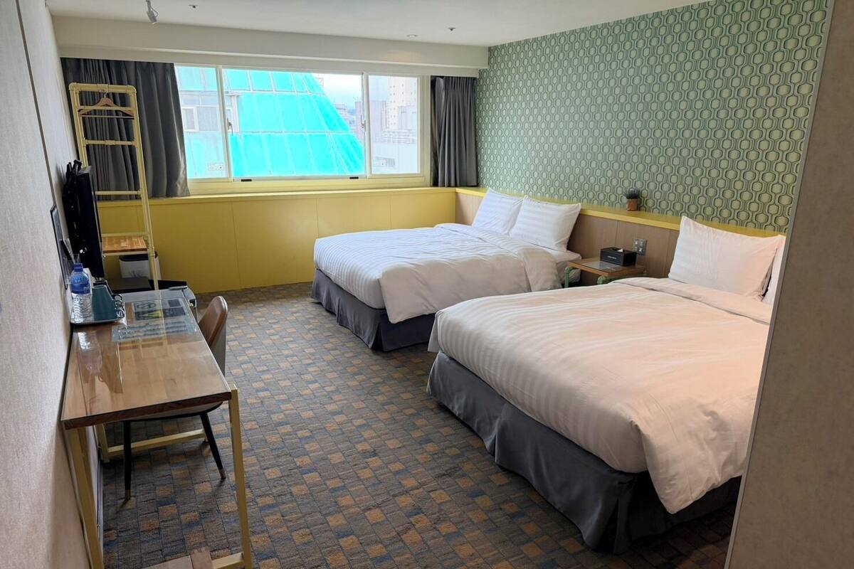 A hotel room with two beds, a desk, a chair, a small table, a window with curtains, and a patterned wall.