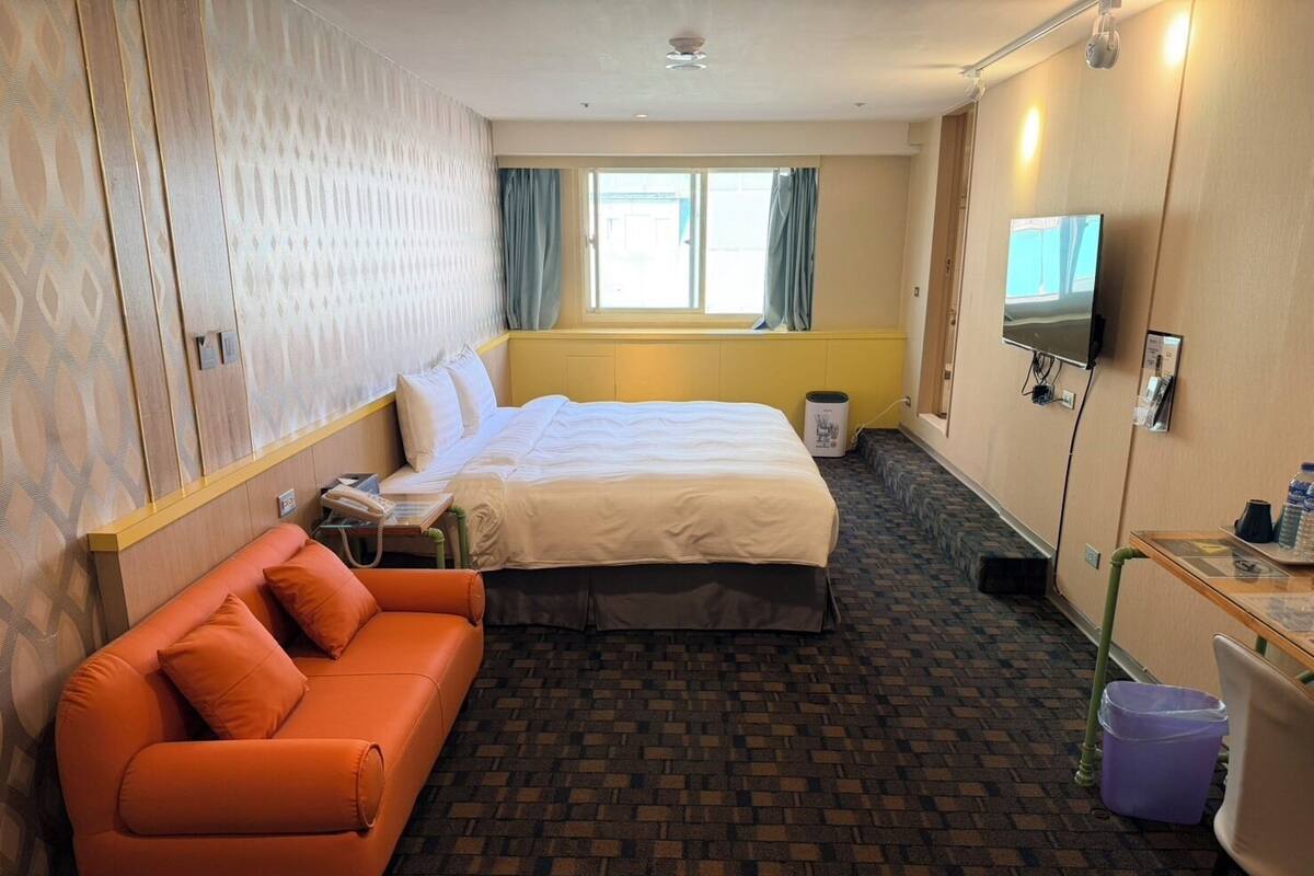 A hotel room with a bed, orange sofa, TV, and a window with curtains.