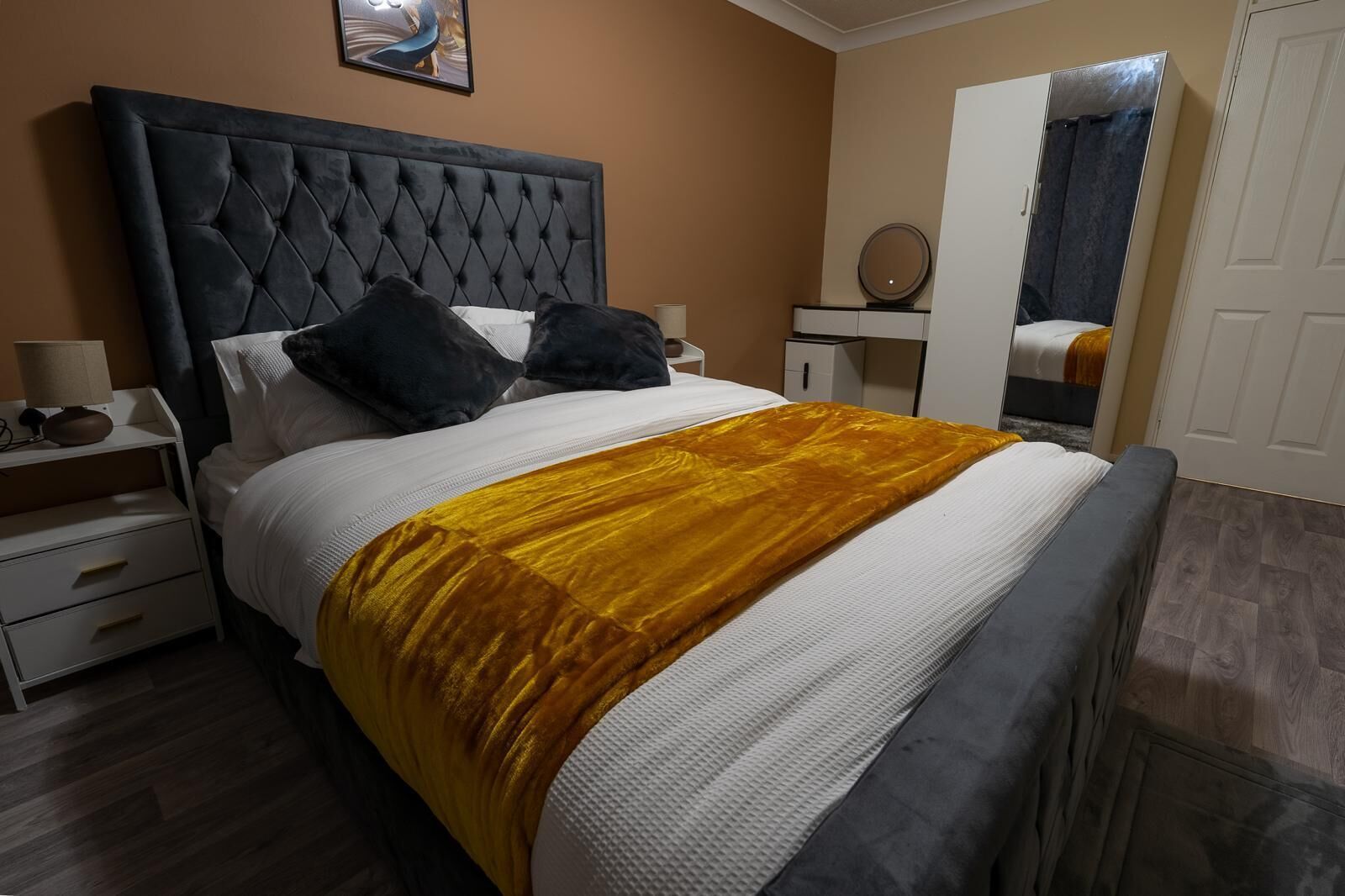 A bedroom with a tufted headboard, a bed with white and dark grey bedding, a mustard-colored throw blanket, a bedside table with a lamp, and a mirror on the wall.
