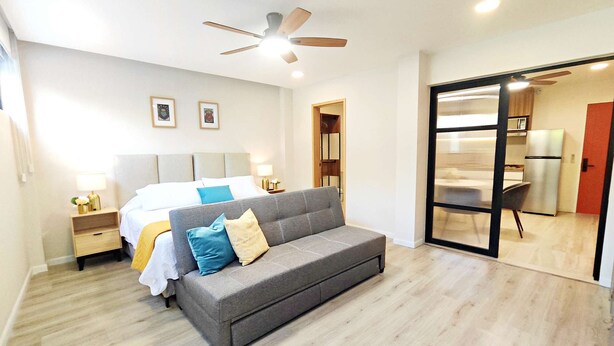 A modern bedroom with a grey sofa, a bed with white and blue bedding, a ceiling fan, and a kitchenette visible through a glass door. - Perfect for Digital Nomads | Studio in Condesa (Mexico City)