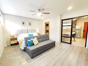 A modern bedroom with a grey sofa, a bed with white and blue bedding, a ceiling fan, and a kitchenette visible through a glass door. - Coco 5 · 30 m² suite with terrace in Condesa · 1 bath (Mexico City)