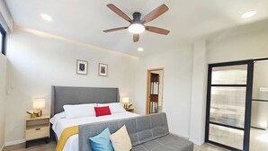 A bedroom with a bed, a grey sofa, a ceiling fan, bedside lamps, and a large window. - Stay in Condesa | New, Cozy & Full of Charm (Mexico City)