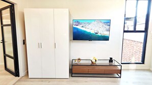 A room with a white wardrobe, a wooden TV stand with a flat-screen TV displaying a coastal scene, and a large window with a view of a brick wall. - Stay in Condesa | New, Cozy & Full of Charm (Mexico City)