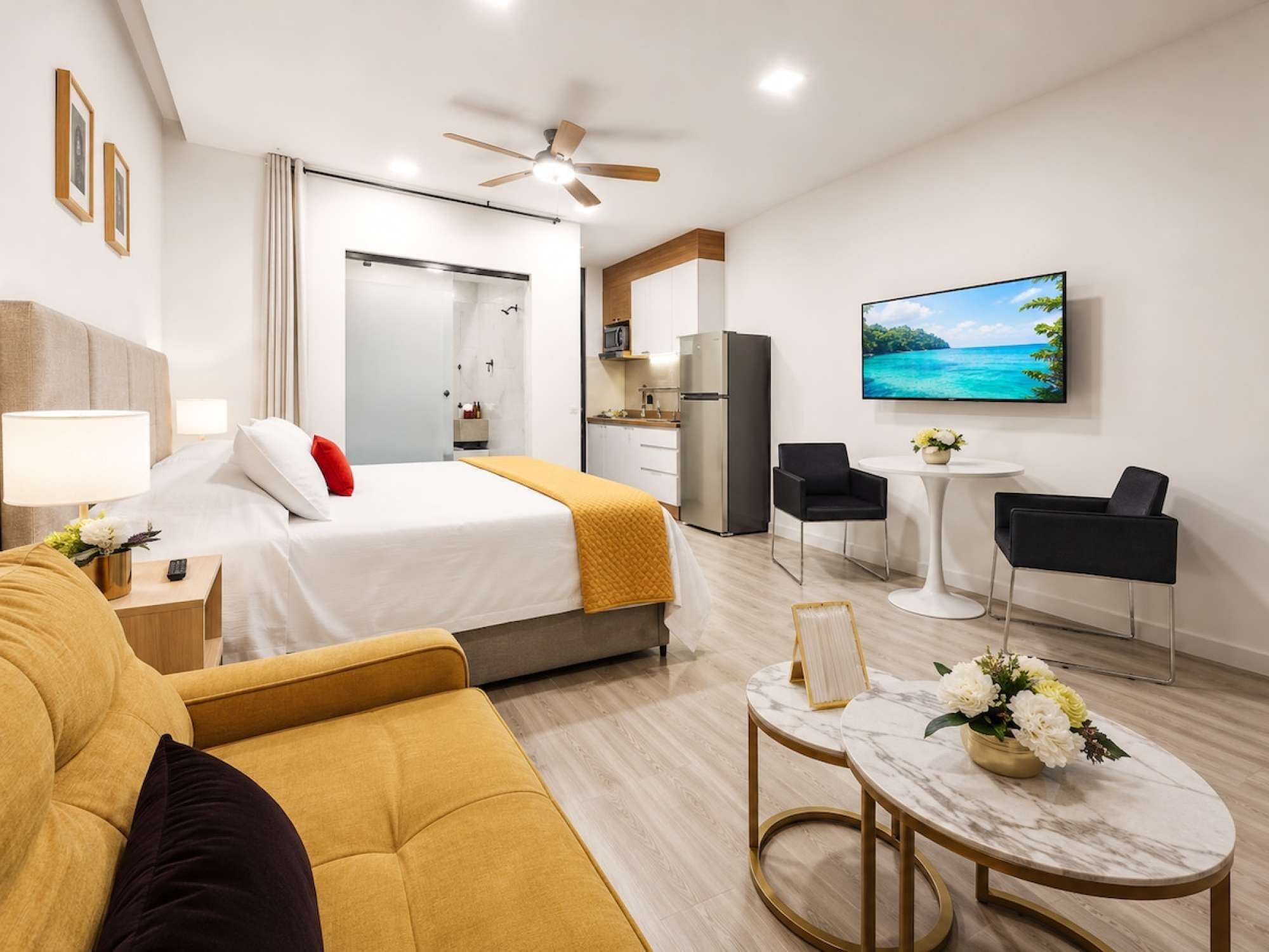 A modern hotel room with a bed, sofa, dining area, and kitchen.
