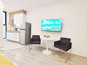 Cocina moderna con armarios blancos, un refrigerador y un microondas. Cuenta con una zona de comedor con dos sillas y una mesa pequeña, y un televisor grande montado en la pared mostrando un paisaje.