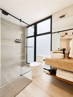 A modern bathroom with a glass shower enclosure, a toilet, a sink with a mirror, and a window providing natural light. - Coco 4 · 20 m² studio with terrace in Condesa · 1 bath (Mexico City)