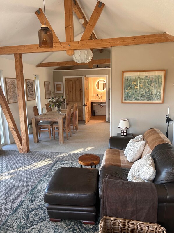 A spacious living room with a wooden ceiling, a leather sofa, a dining area with wooden chairs, and a kitchen visible through an open door. - Waterside Cabin | Cavan Lakes | Hot Tub (CN)