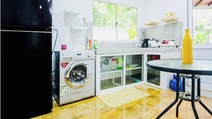 Une cuisine compacte équipée d’un lave-linge, d’une armoire vitrée, d’une petite table et d’une fenêtre donnant sur un espace verdoyant.