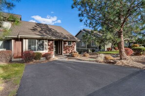 A single-story house with a front entrance, a driveway, and a well-maintained lawn with various shrubs and a pine tree. - Eagle Crest Village Loop 10840 | Tranquil Retreat (Redmond)