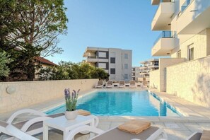 Une piscine entourée de chaises longues et agrémentée d’une plante en pot, le tout bordé d’un mur de pierre et surplombé par des bâtiments modernes.