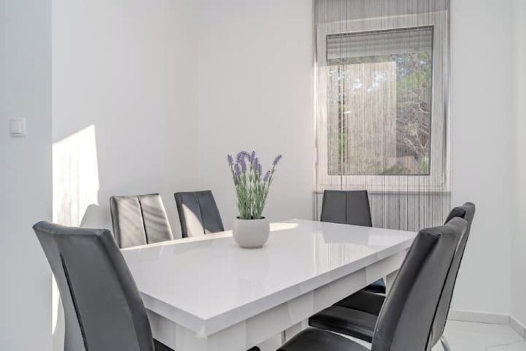 Uma sala de jantar moderna com uma mesa branca, seis cadeiras e um vaso de planta.