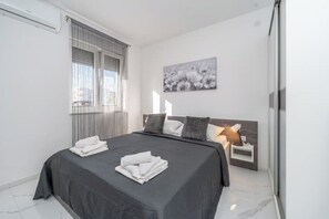 Quarto moderno com uma cama grande, uma mesa de cabeceira com abajur e uma janela com persianas.