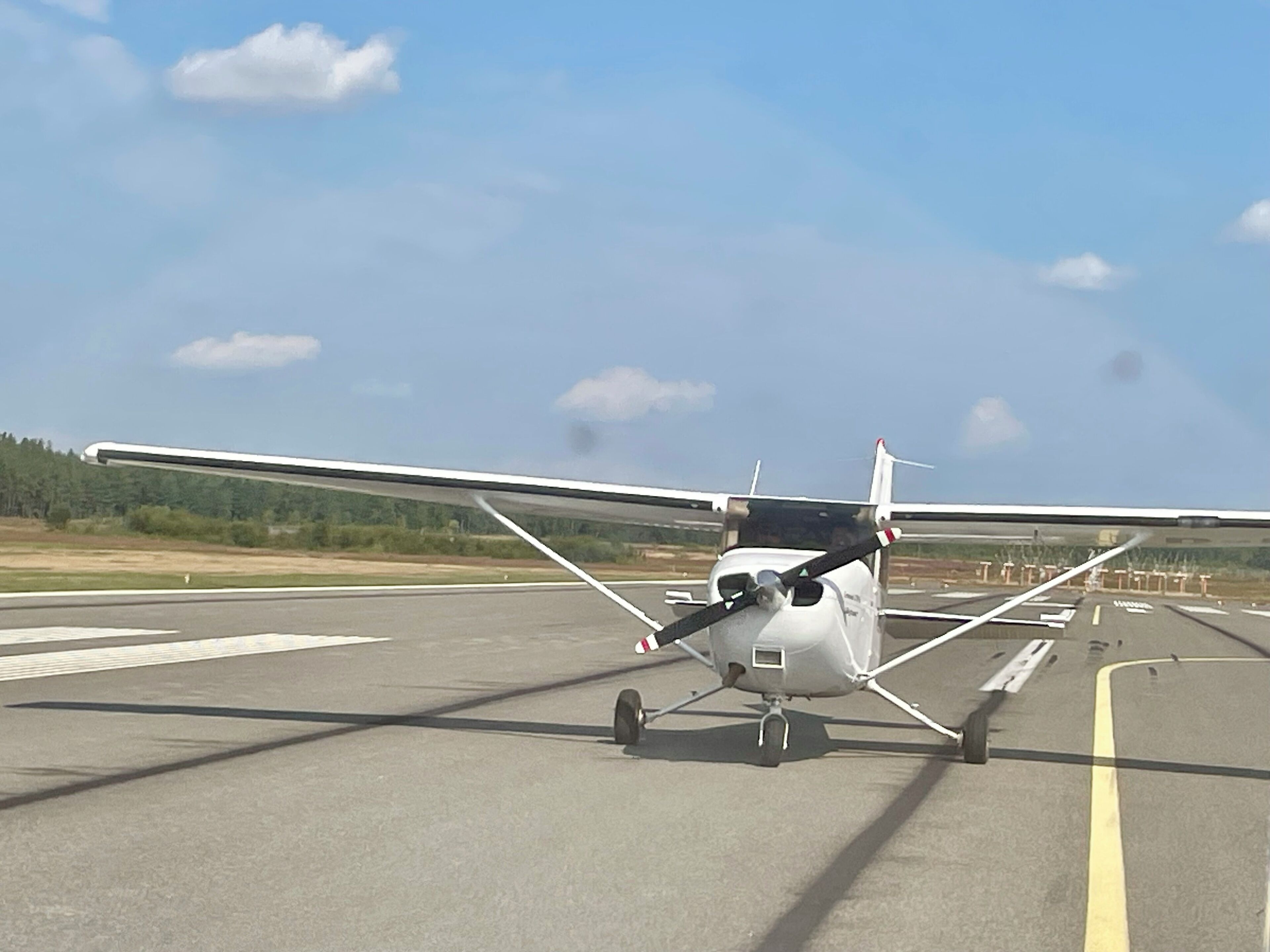 Ein kleines, weißes Flugzeug auf einer Startbahn bei klarem Himmel und einem Wald im Hintergrund.