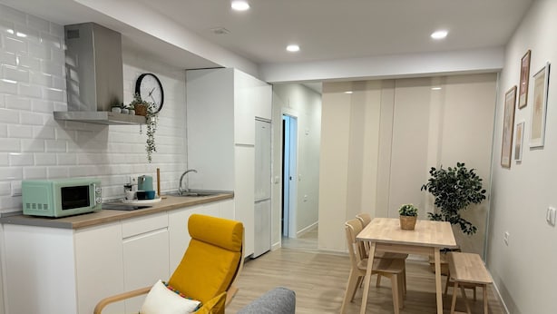 Eine moderne Küche mit weißen Schränken, einer Mikrowelle und einem Spülbecken. Im Wohnbereich befindet sich ein gelber Sessel und ein graues Sofa. Im Hintergrund ist ein Holzesstisch mit Stühlen zu sehen.