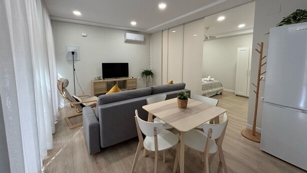 Ein modernes Wohnzimmer mit einer Couch, einem Esstisch und Stühlen. Ein Fernseher auf einem Schrank, eine Pflanze und ein weißer Kühlschrank sind ebenfalls vorhanden.