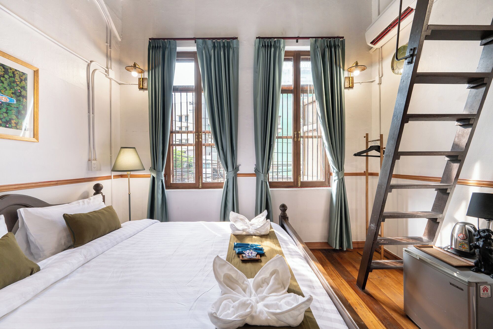 Habitació d'hotel amb un llit, un escriptori, una escala i dues finestres amb cortines.