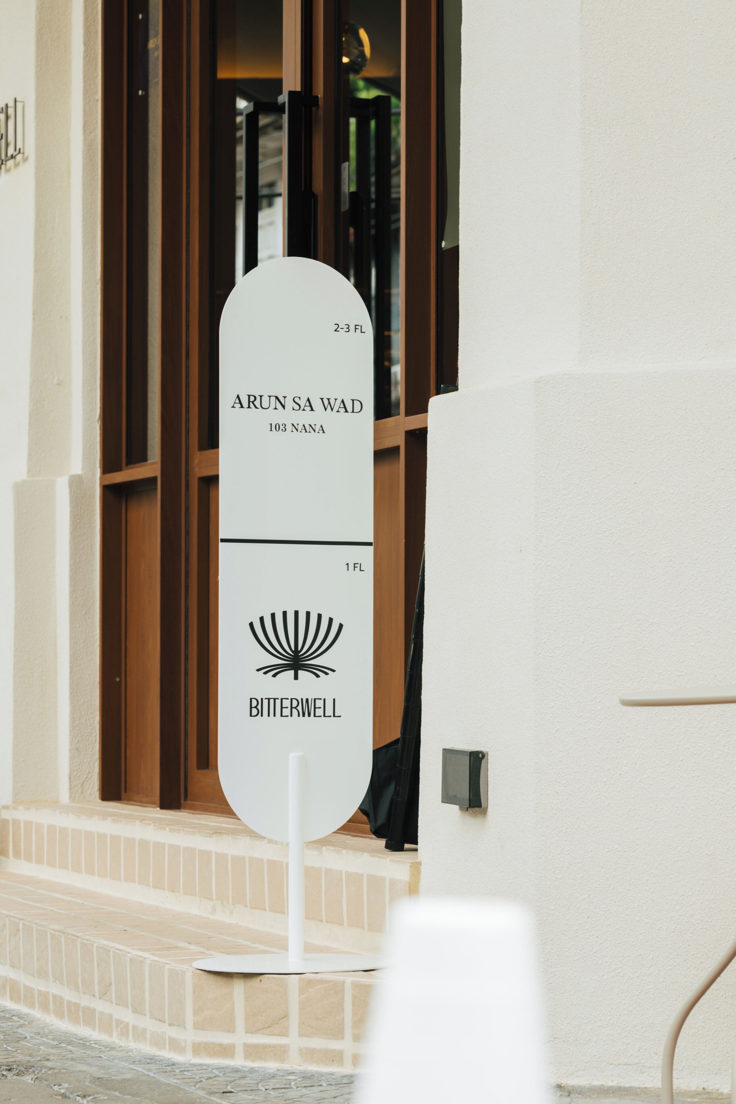 A sign with 'ARUN SA WAD 103 NANA' and 'BITTERWELL' on it, placed outside a building with a wooden door and steps.