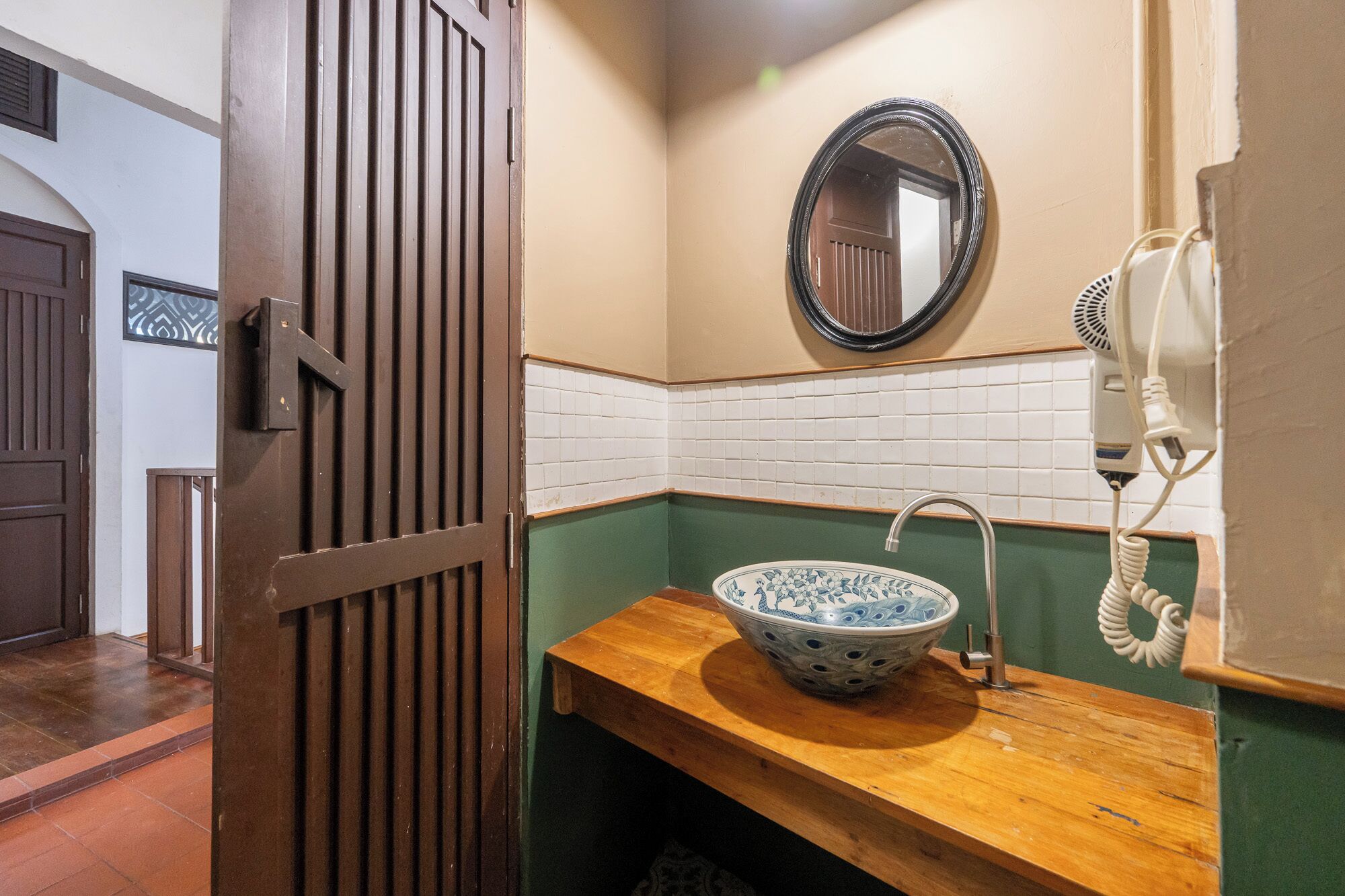 Ein Badezimmer mit einem Holzlagerflächenmöbel, einem weißen Waschbecken mit blumigem, blauem Design, einem runden Spiegel und einem Fön an der Wand.