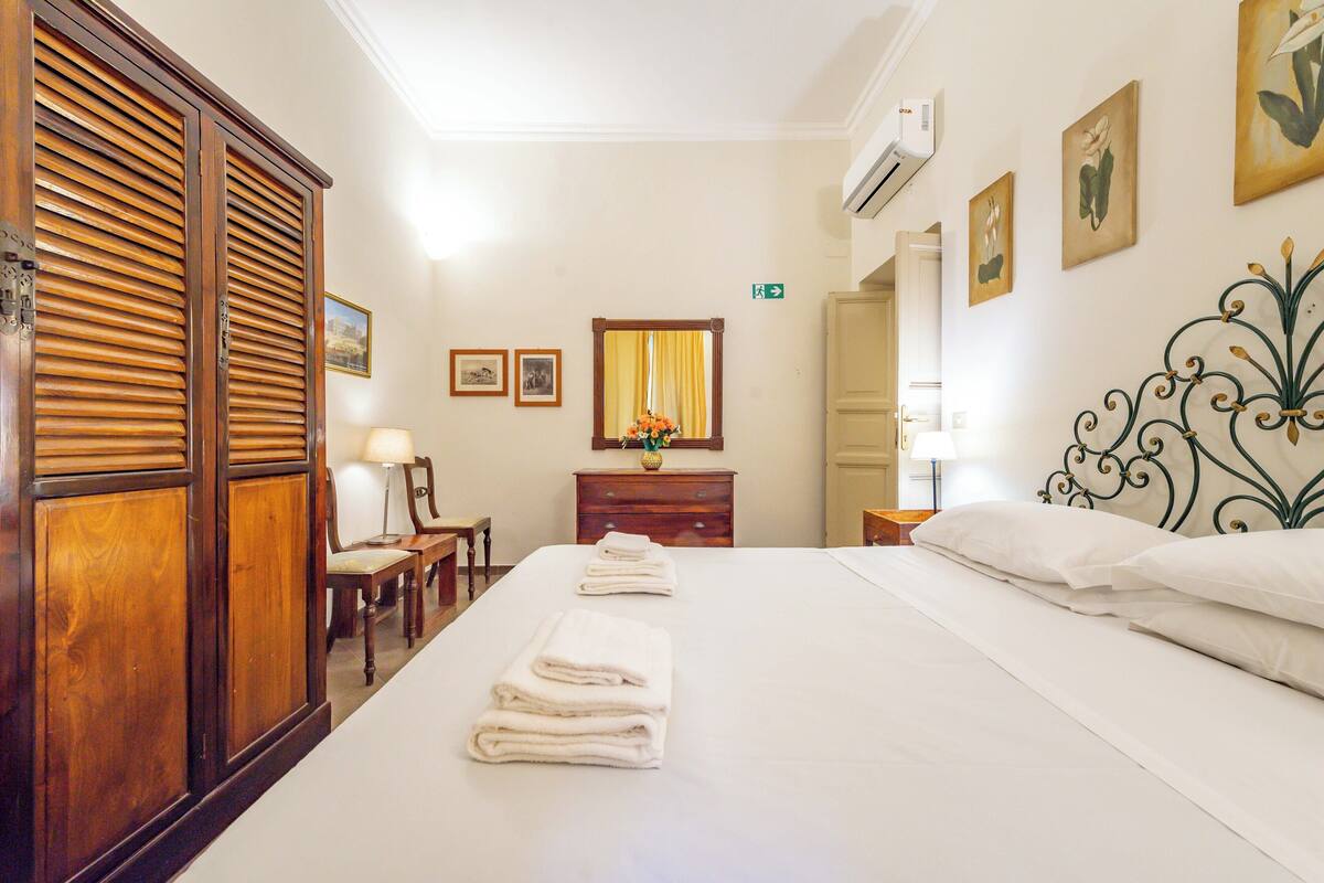A hotel room with a large bed, wooden wardrobe, a desk with a chair, a mirror, and a small table with a lamp.