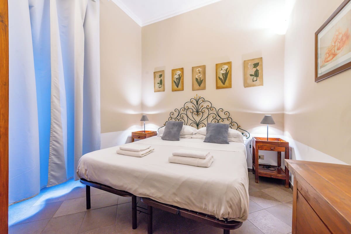 A neatly made bed with pillows and folded towels, a bedside table with a lamp, and wall art above the bed.