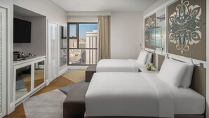Quarto de hotel com duas camas, televisão, secretária com espelho e uma janela grande com vista para a cidade.