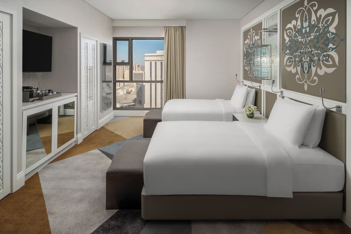 A hotel room with two beds, a TV, a desk with a mirror, and a large window with a city view.