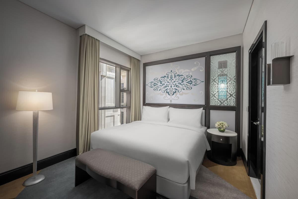 A hotel room with a bed, a bedside table with a lamp, a window with curtains, and a decorative wall piece.