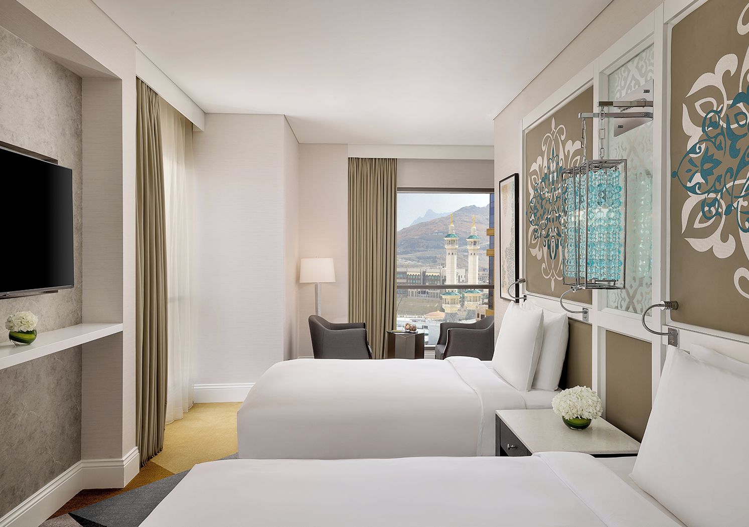 Habitació d'hotel amb dos llits, una televisió, una làmpada, vista a una mesquita i una peça decorativa a la paret.