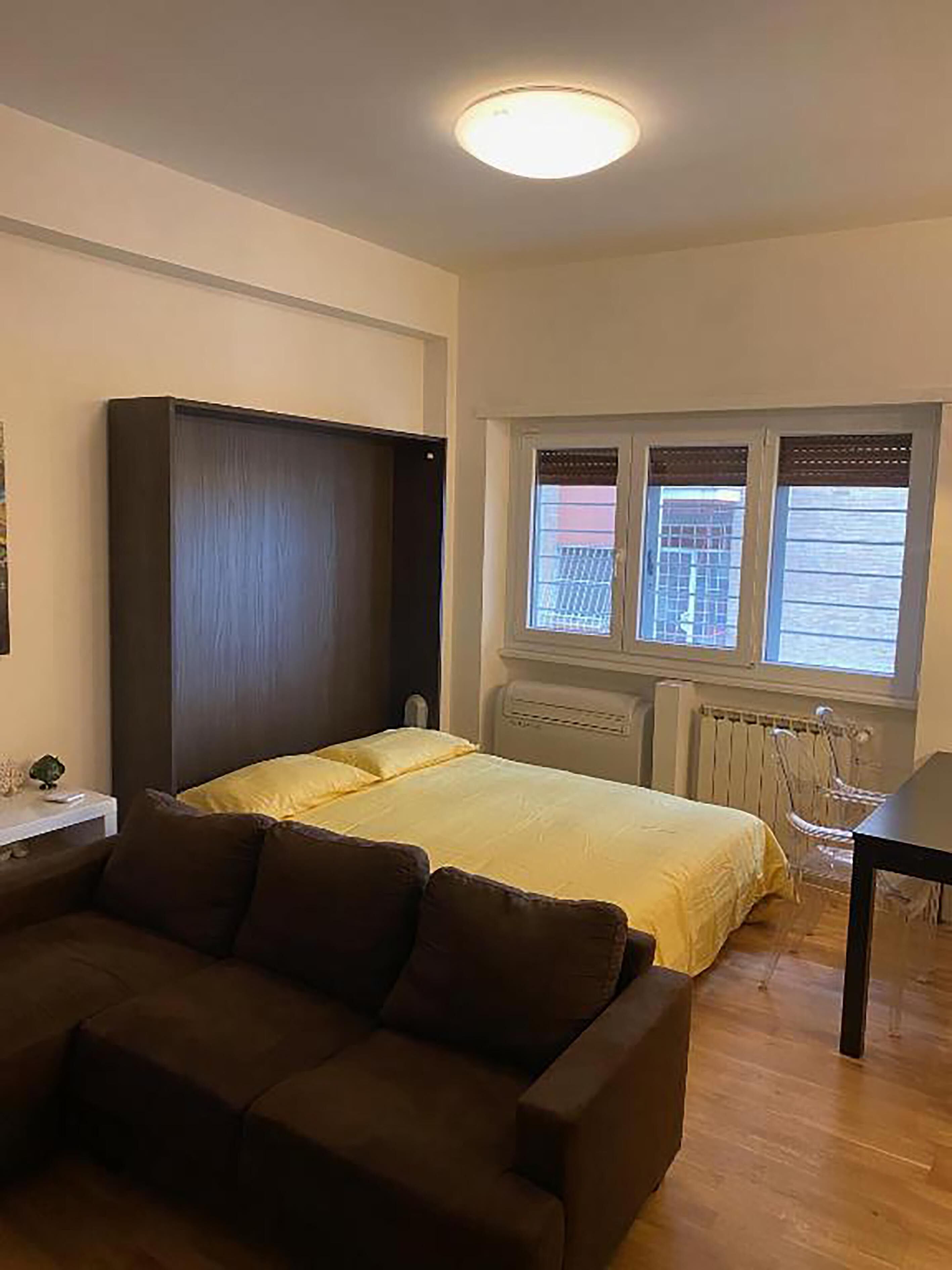 A bedroom with a bed, a dark-colored sofa, a small table, and a window with blinds.