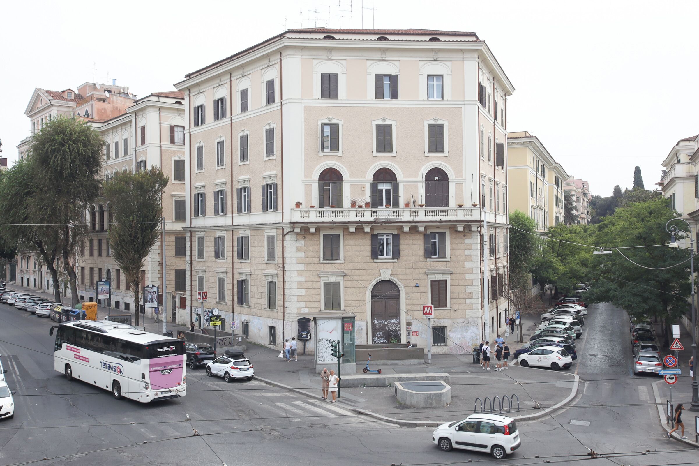 A multi-story building with a central entrance, a bus stop, and a row of parked cars along the street.