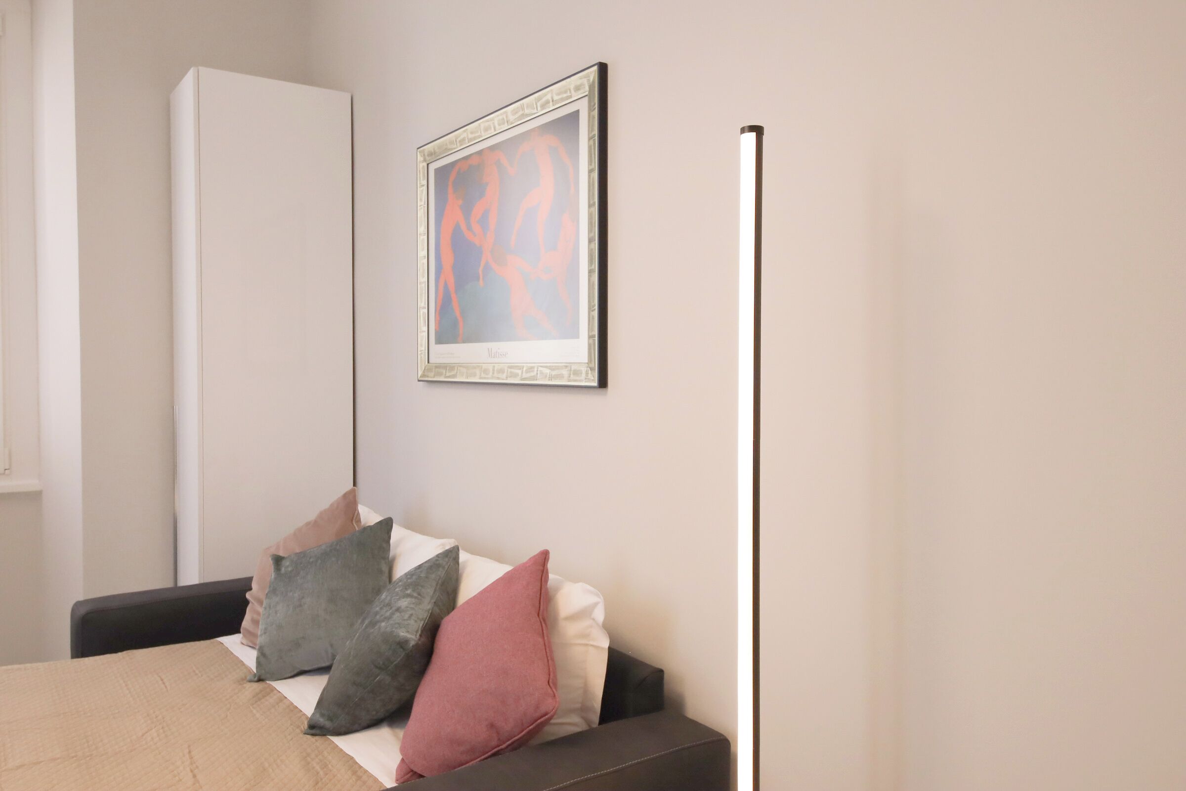 A bedroom with a bed, pillows, a bedside table, a wardrobe, and a framed artwork on the wall.