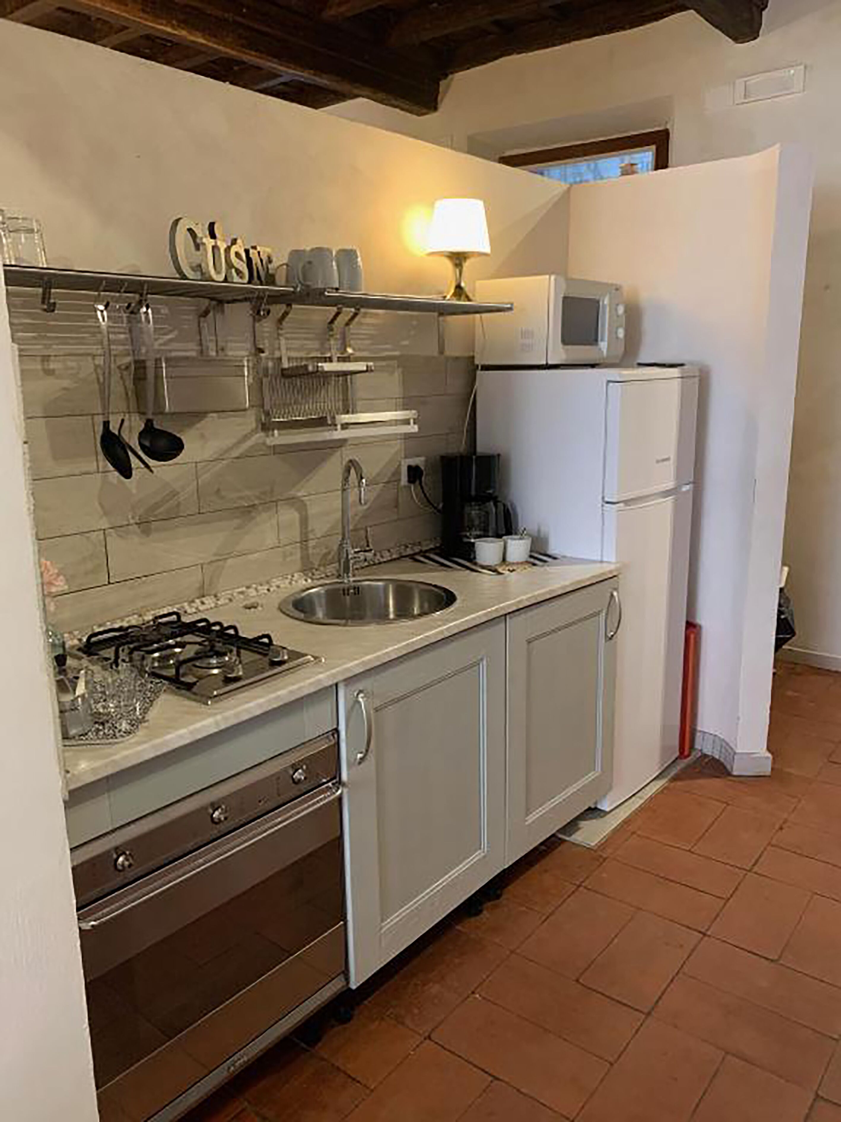 Une cuisine compacte équipée d’une cuisinière à gaz, d’un évier et de meubles blancs. On peut également apercevoir un four à micro-ondes et un petit réfrigérateur.