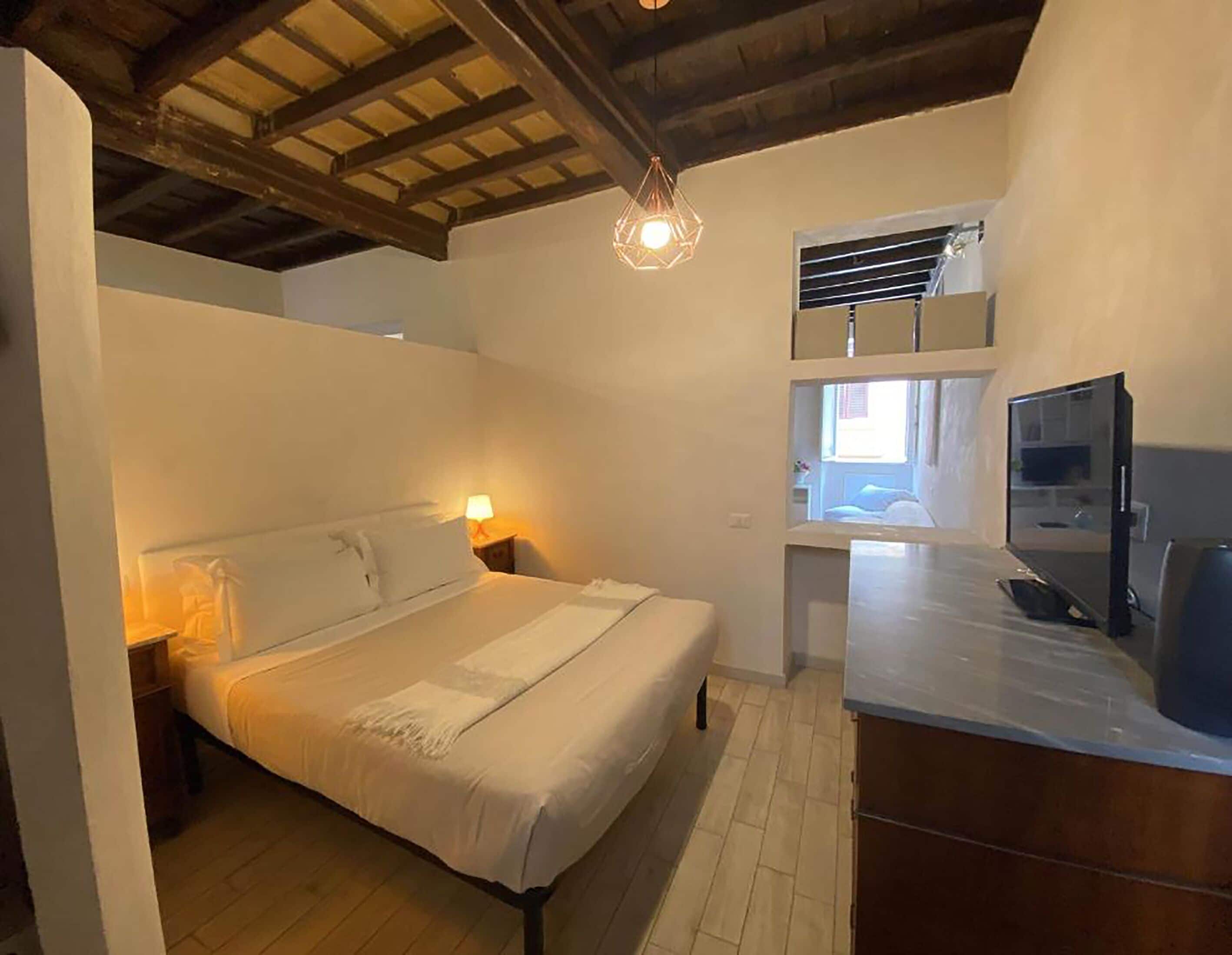 Ein Schlafzimmer mit Holzboden, einem Bett mit weißen Leinen, einem Nachttisch mit Lampe, einem kleinen Fenster und einer Küchenzeile.