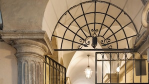 An arched doorway with a metal gate, a column, and a hallway leading to a room with a ceiling light. - Elegant Apartment Minelli Navona by Ghor (Rome)