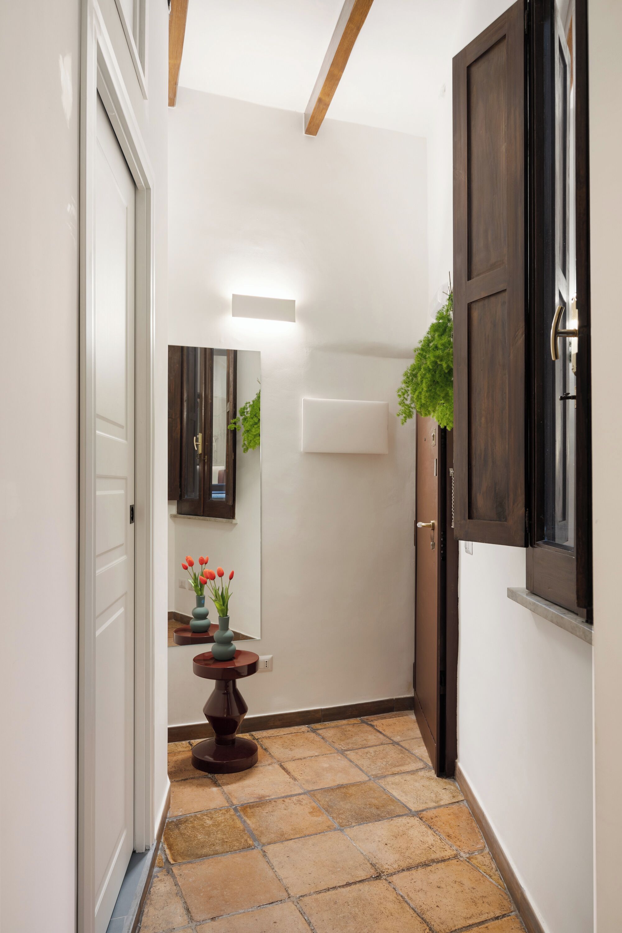 A narrow hallway with a tiled floor, a wooden door, a small table with a vase of flowers, and a mirror on the wall.