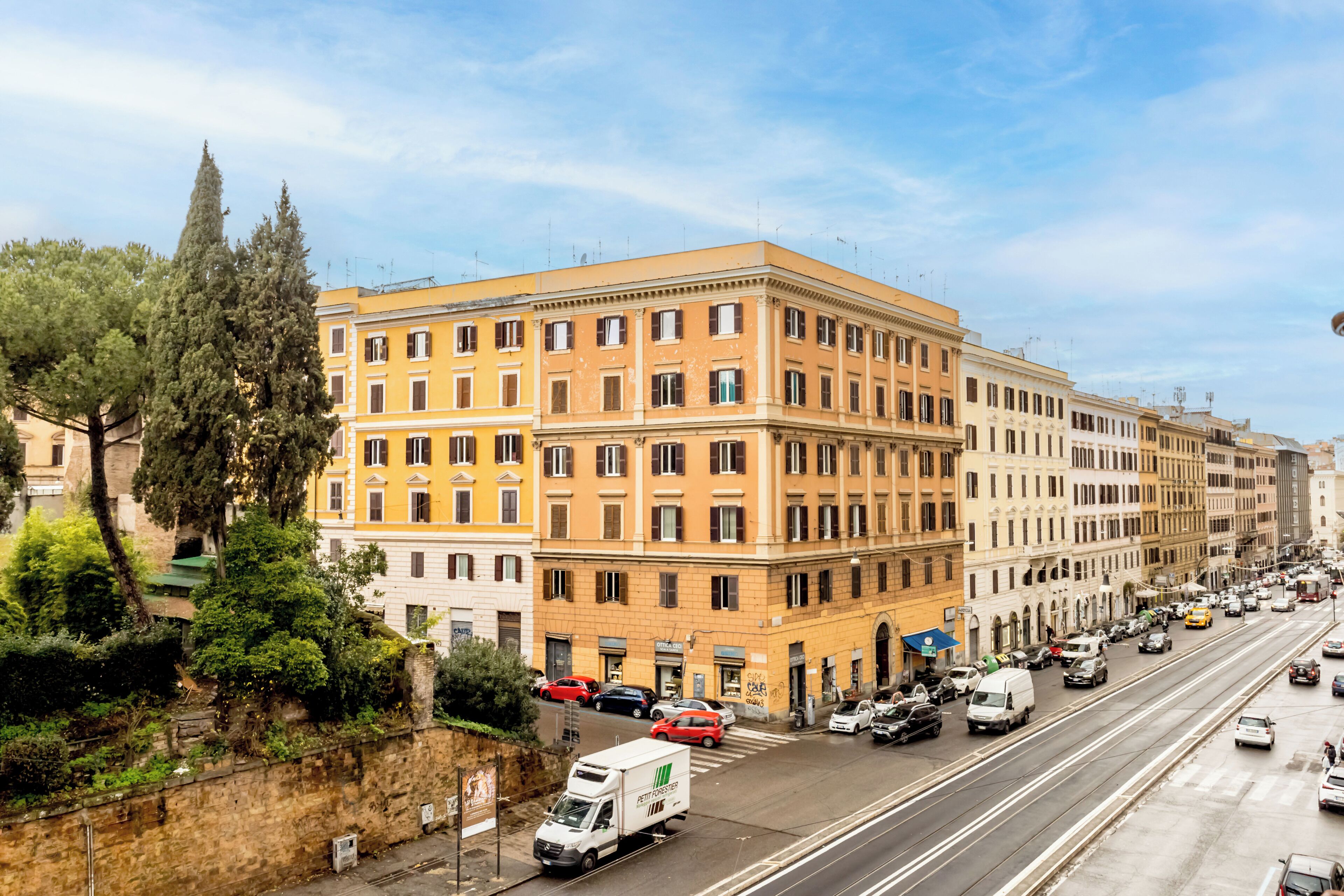 Ein mehrstöckiges Gebäude mit einem Hauseingang in der Ecke, eine Straße davor mit Autos und einem weißen Lieferwagen, sowie Bäume auf der linken Seite.