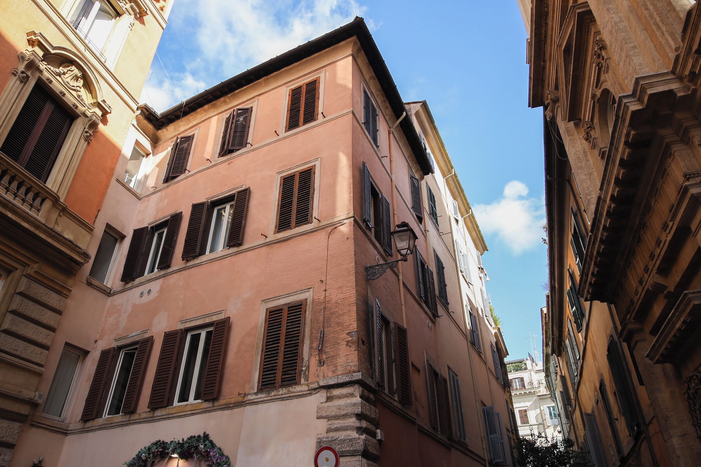 A narrow street with historic buildings, including a pink building with shutters and a visible no parking sign.