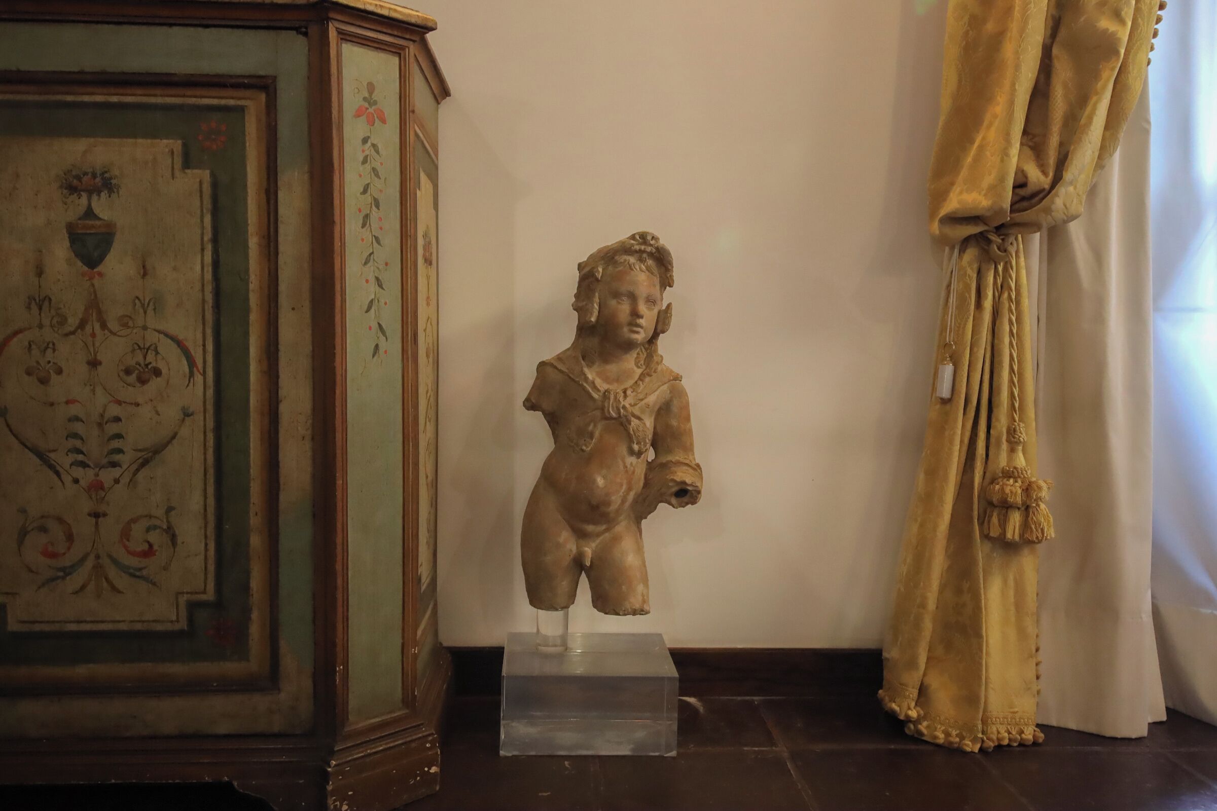 Una escultura sobre un pedestal transparente, un aparador decorativo con motivos florales y una cortina dorada.
