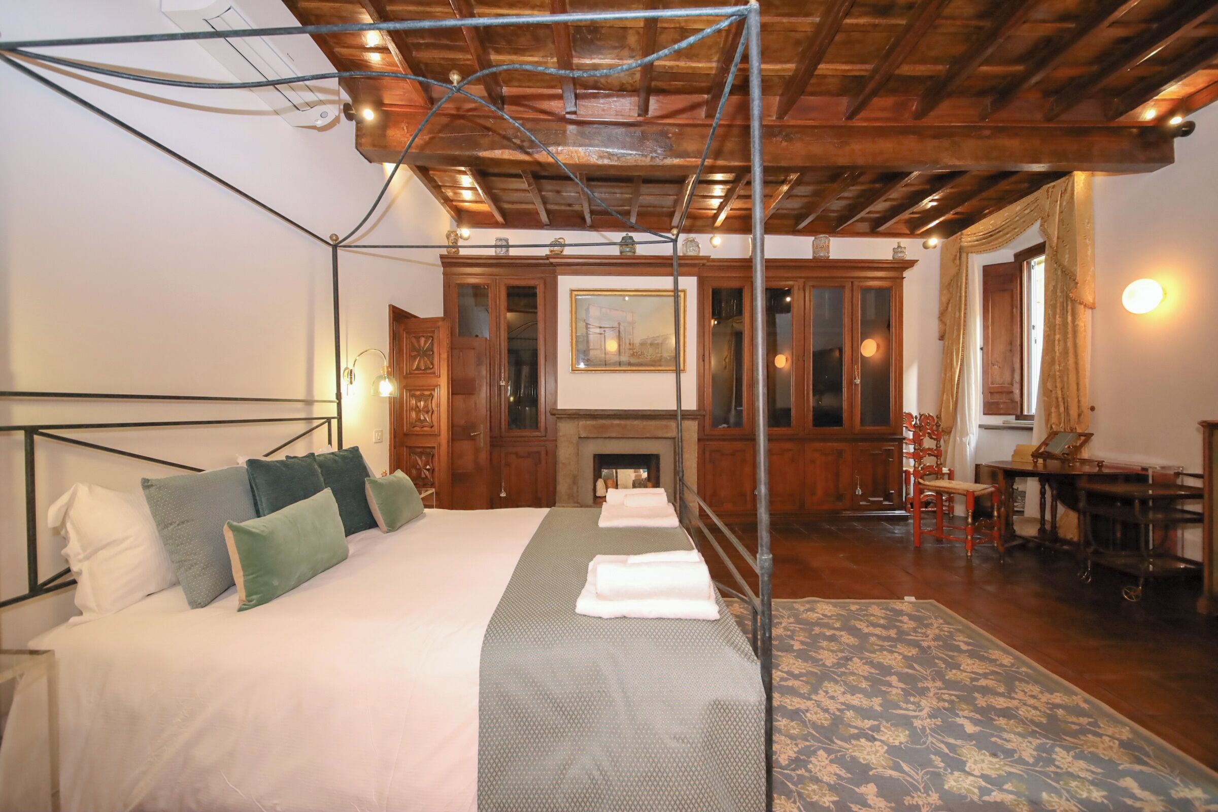 A bedroom with a wooden ceiling, a bed with white and green bedding, a fireplace, and a dining area with wooden furniture.