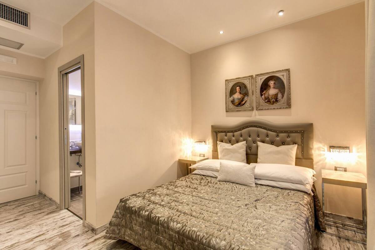 Un dormitorio con una cama grande, dos mesitas de noche con lámparas, una puerta que da a otra habitación y dos cuadros en la pared.