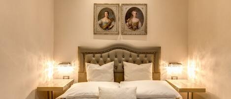 Une chambre à coucher avec une tête de lit capitonnée, deux tables de chevet avec des lampes et deux portraits encadrés accrochés au mur.