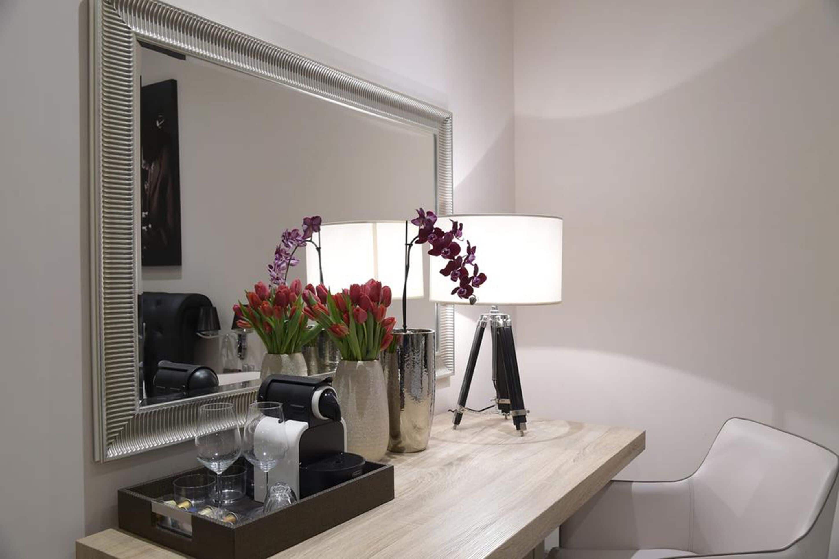 A hotel bathroom with a large mirror, a vanity with a tray of toiletries, a lamp, and a vase of flowers.