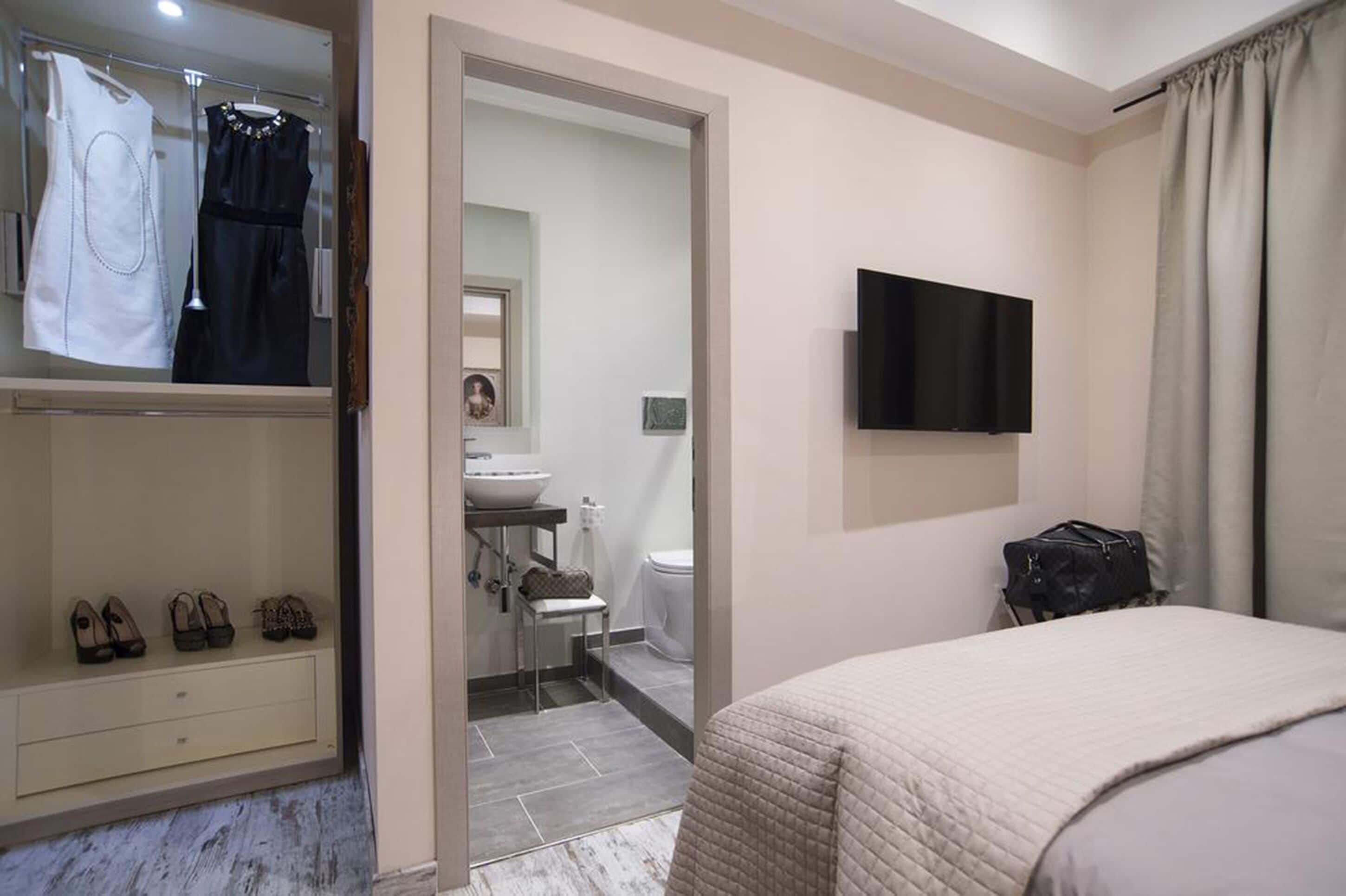 Une chambre à coucher avec un lit, un téléviseur fixé au mur, une armoire remplie de chaussures et une salle de bain visible à travers une porte ouverte.
