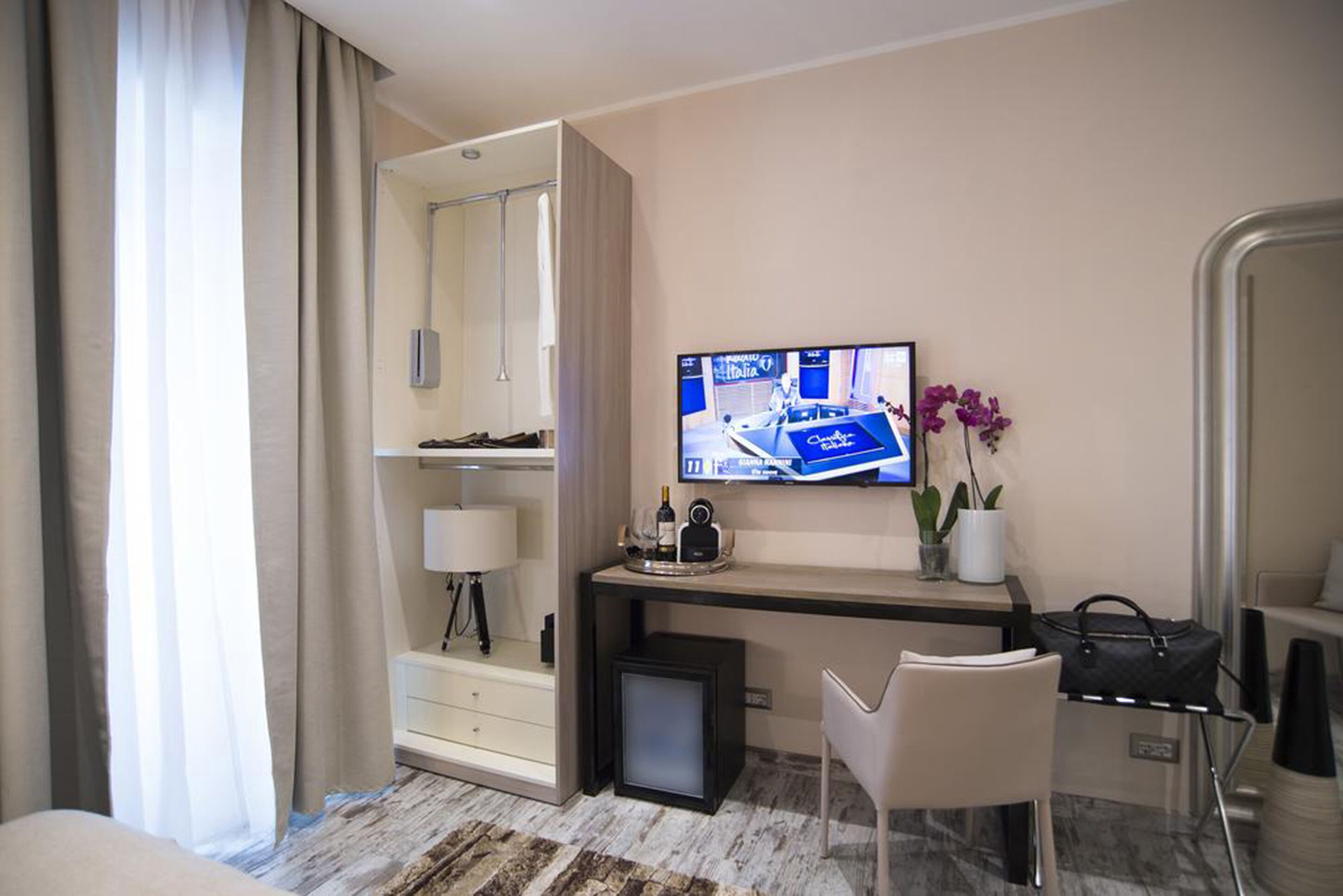 Quarto de hotel com cama, escrivaninha com televisão, cadeira, abajur, mesinha com garrafa e xícara, e uma planta.