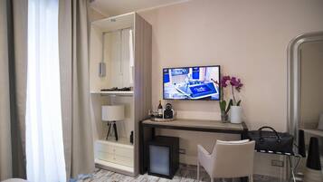 Quarto de hotel com cama, escrivaninha com televisão, cadeira, abajur, mesinha com garrafa e xícara, e uma planta.