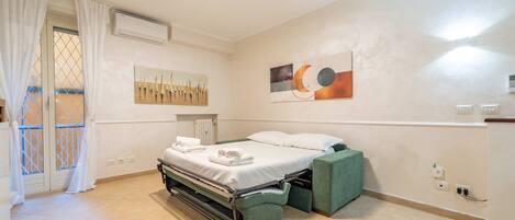 A hotel room with a bed, a green chair, a window with curtains, and artwork on the walls.